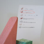 Block Woman reviews a hand-written list of potential charitable donations. There is a pile of cash underneath the list.