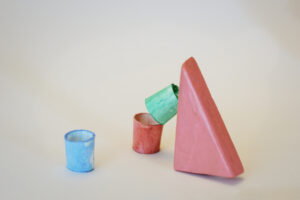 Block Woman surveys three pastel colored buckets: blue, red and green. The green bucket is balanced on top of the red bucket and is tilted against Block Woman.