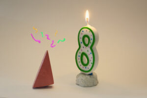 Block Woman with colored squiggles symbolizing incredulity gazes at a lit number 8 birthday candle atop a miniature white birthday cake.