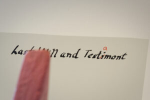 Block Woman stands in front of a white sheet of paper with the title written in old script that says Last Will and Testimont. The letter i in misspelled Testimont is crossed through in red and a letter a is written above.