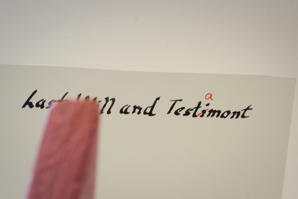 Block Woman stands in front of a white sheet of paper with the title written in old script that says Last Will and Testimont. The letter i in misspelled Testimont is crossed through in red and a letter a is written above.