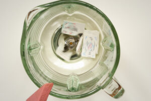 Block Woman peers over the edge of a clear blender, looking down on an ice cube and the stainless steel blender blade.