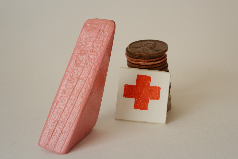 Block Woman stands next to a stack of copper pennies with a white square with a red cross symbol leaning against the pennies.