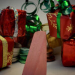 Block Woman stands in front of a pile of boxes gift-wrapped in shiny red paper and tied with green and gold ribbons.