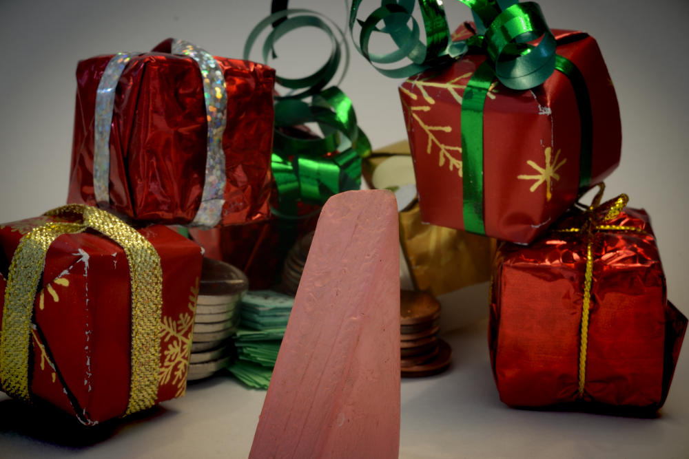 Block Woman stands in front of a pile of boxes gift-wrapped in shiny red paper and tied with green and gold ribbons.